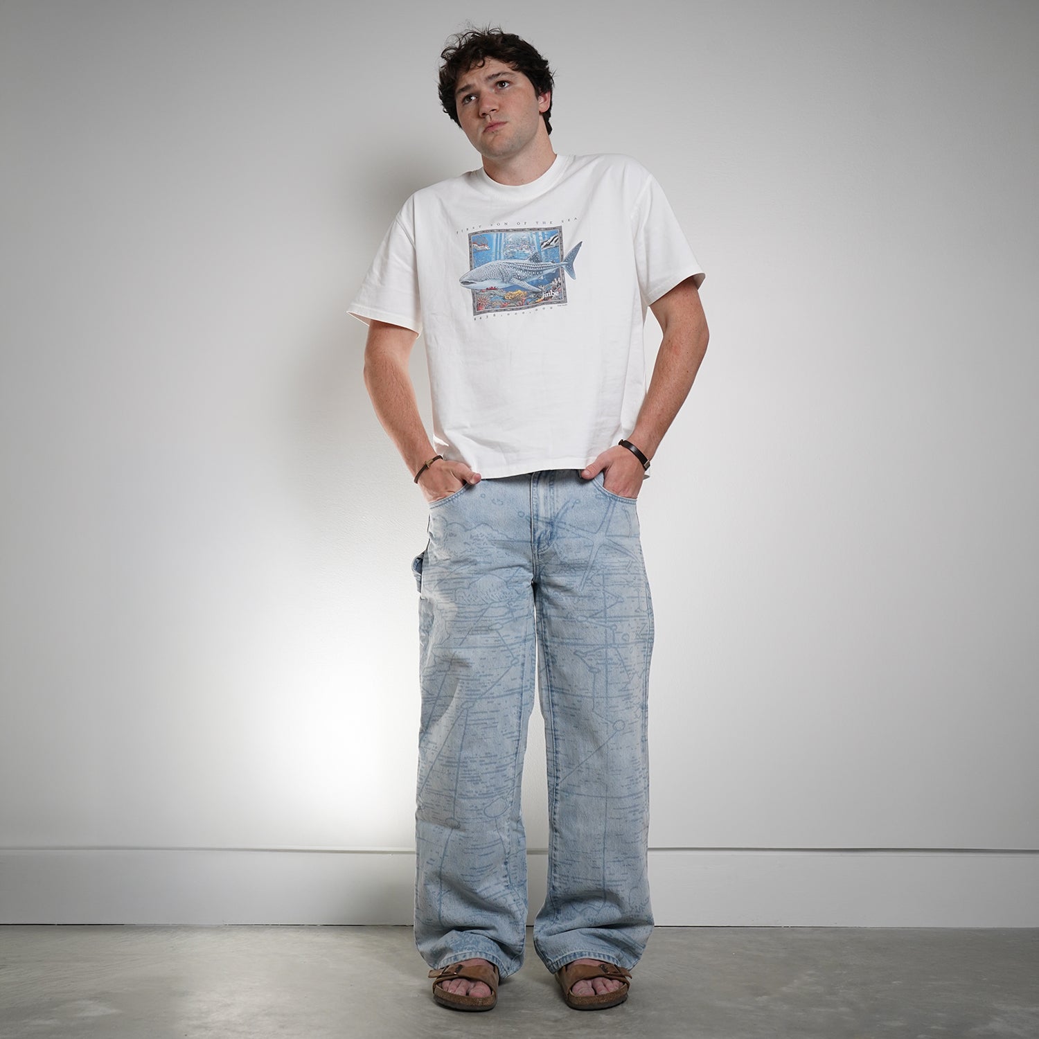 Man wearing a white t-shirt with a graphic design and blue jeans against a plain background