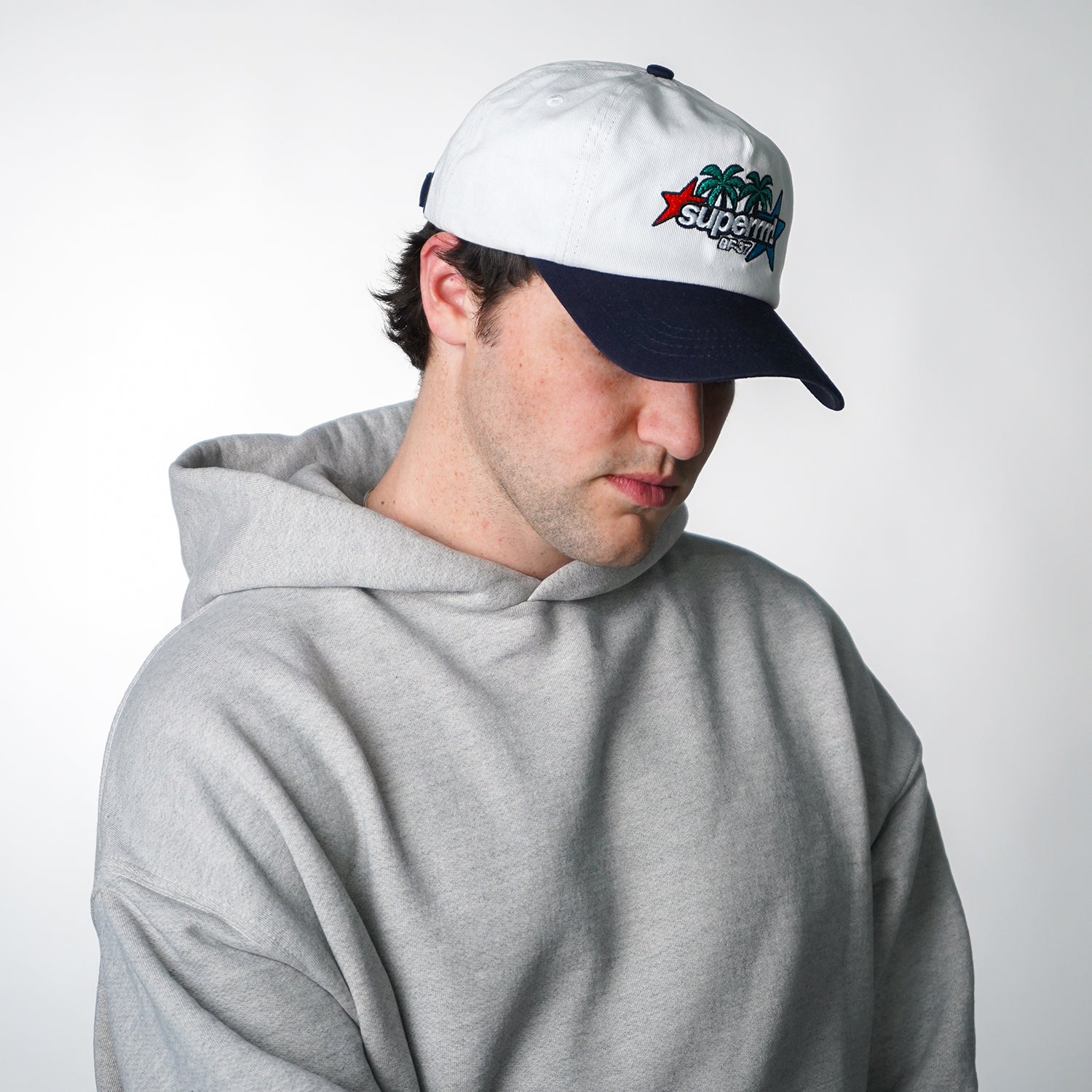 Person wearing a gray hoodie and a white and navy cap with a logo on a light gray background