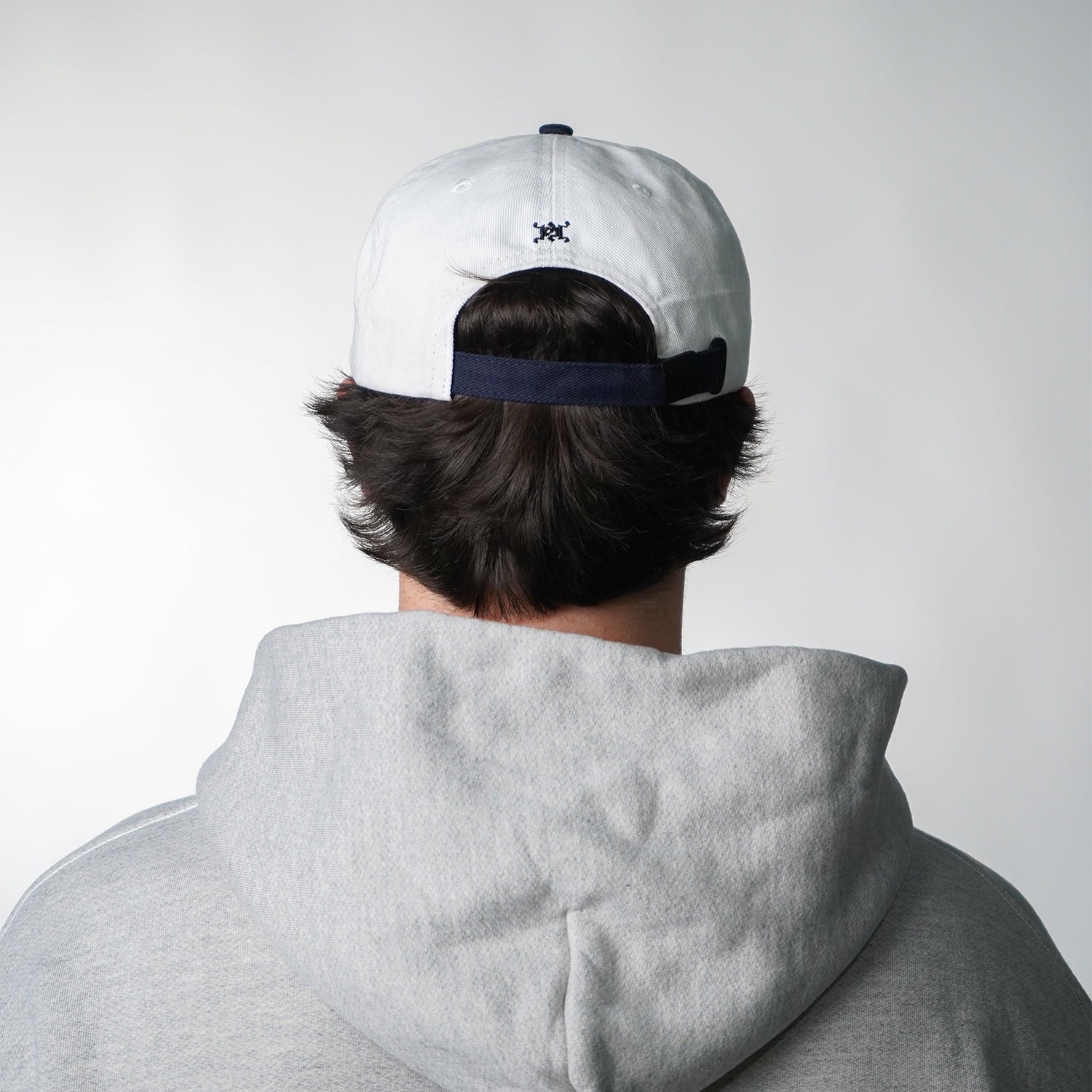 Person wearing a white cap with a black logo and a gray hoodie on a plain background