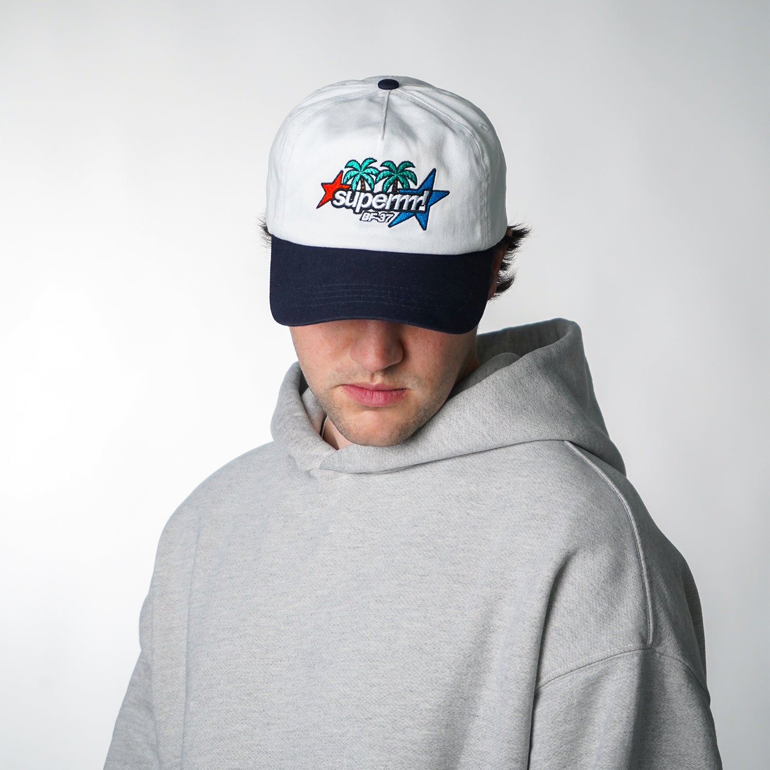 Person wearing a white cap with a colorful logo and a gray hoodie on a light gray background